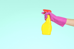 A bright yellow spray bottle is being held aloft by a hand wearing a bright pink cleaning glove. 