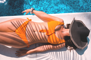 A woman in an orange bikini lies beside a pool with a sun hat covering her head, and the sun shining on her skin