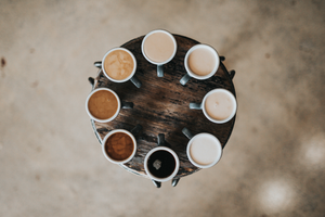 Photographed from above, 8 coffees of darkening colors sit in a circle on a round wooden coffee table. 
